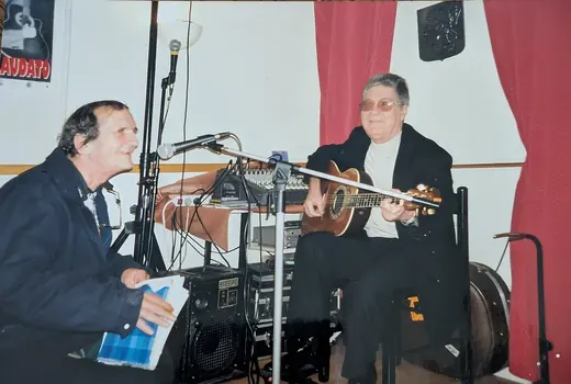 Hubert Tempête, un cahier bleu à la main, et un guitariste, assis devant leurs micros
